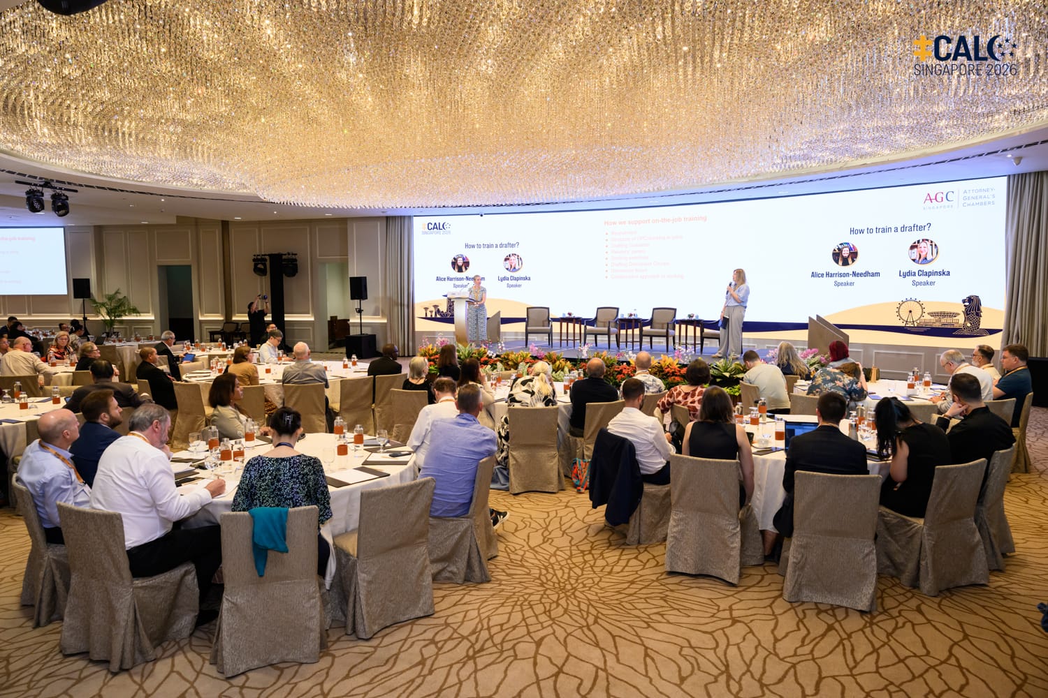 A wide shot of the ballroom at The Fullerton Hotel Singapore during a Conference session, with speakers Alice Harrison-Needham and Lydia Clapinska presenting on stage.
